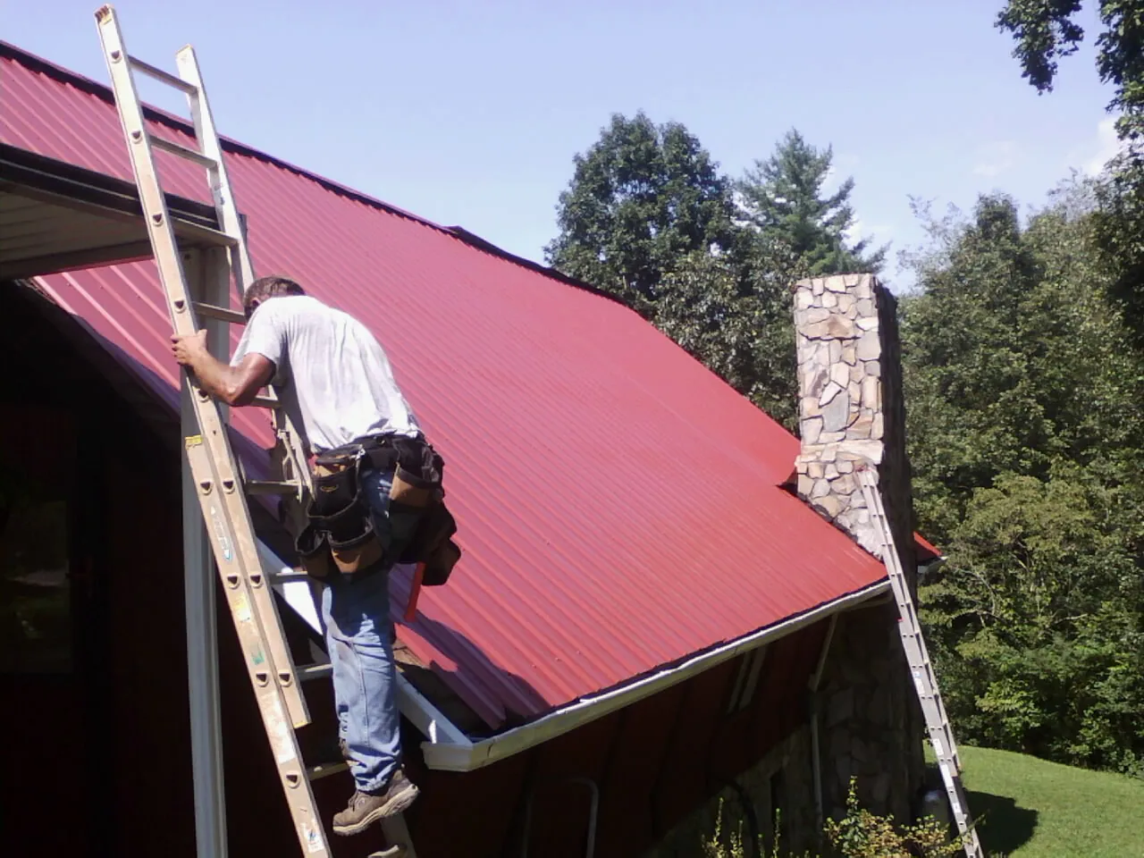 Roofer on a ladder beside a red metal roof and stone chimney — Soffit & Fascia Repair by Roofing Brothers in Cheswick